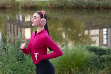Photo of Beautiful woman in headphones running near river outdoors. Space for text