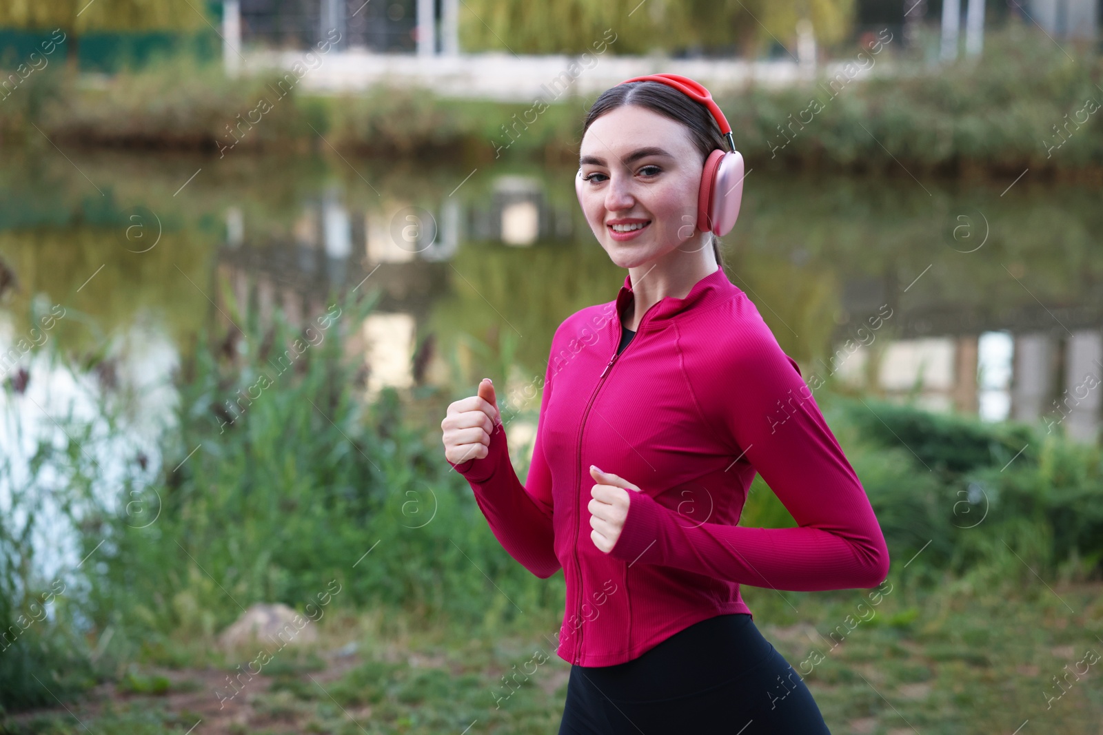 Photo of Smiling woman in headphones running near river outdoors. Space for text