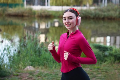 Photo of Smiling woman in headphones running near river outdoors. Space for text