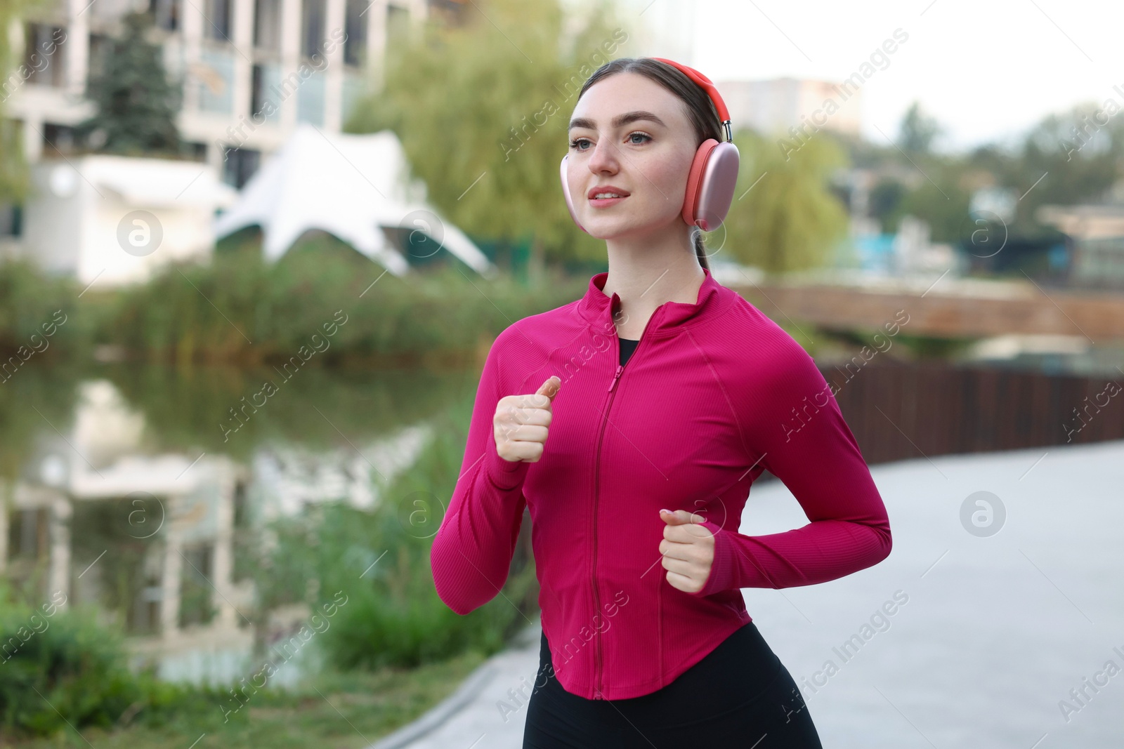 Photo of Smiling woman in headphones running near river outdoors. Space for text