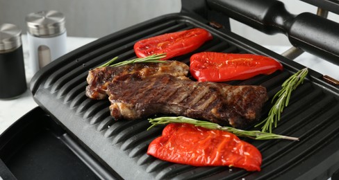 Photo of Electric grill with beef steaks, bell peppers and rosemary on white table, closeup