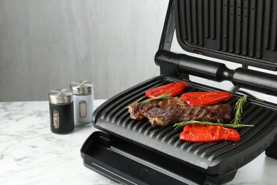Photo of Electric grill with beef steaks, bell peppers and rosemary on white marble table, closeup. Space for text