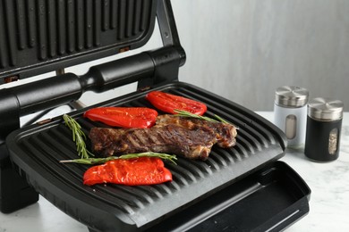 Photo of Electric grill with beef steaks, bell peppers and rosemary on white marble table, closeup