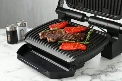 Photo of Electric grill with beef steaks, bell peppers and rosemary on white marble table, closeup
