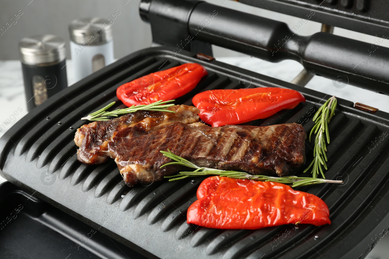 Photo of Electric grill with beef steaks, bell peppers and rosemary on white table, closeup