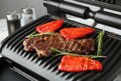 Photo of Electric grill with beef steaks, bell peppers and rosemary on white table, closeup