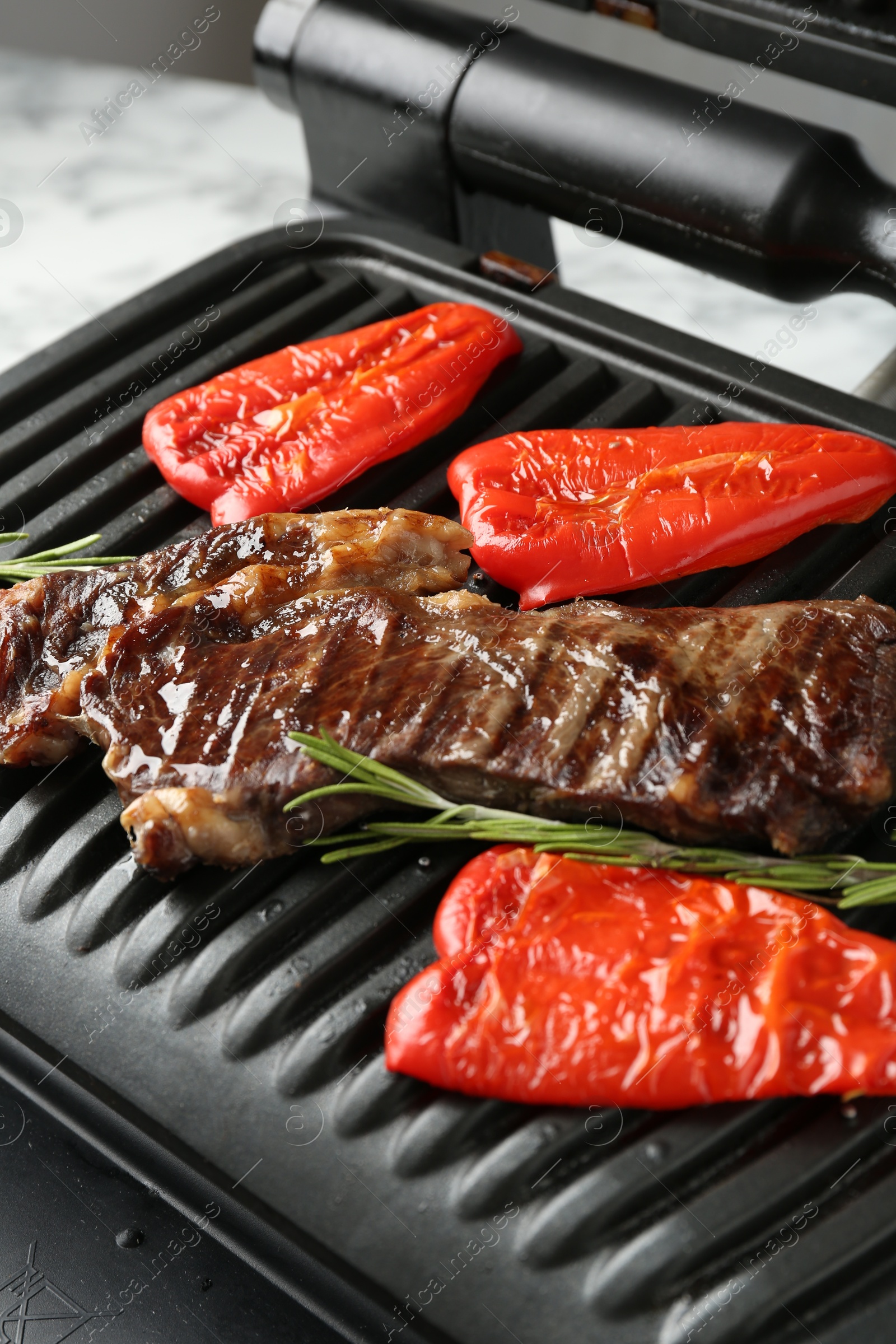 Photo of Electric grill with beef steaks, bell peppers and rosemary on white table, closeup