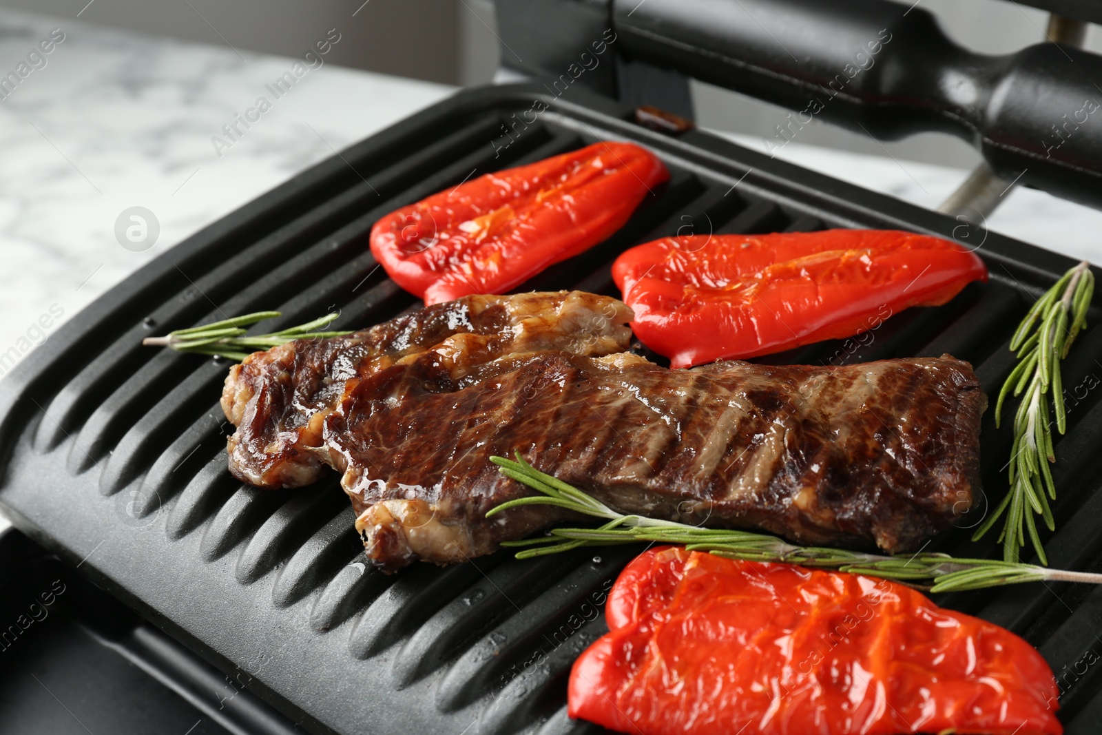 Photo of Electric grill with beef steaks, bell peppers and rosemary on white marble table, closeup