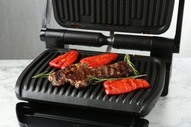Photo of Electric grill with beef steaks, bell peppers and rosemary on white marble table, closeup
