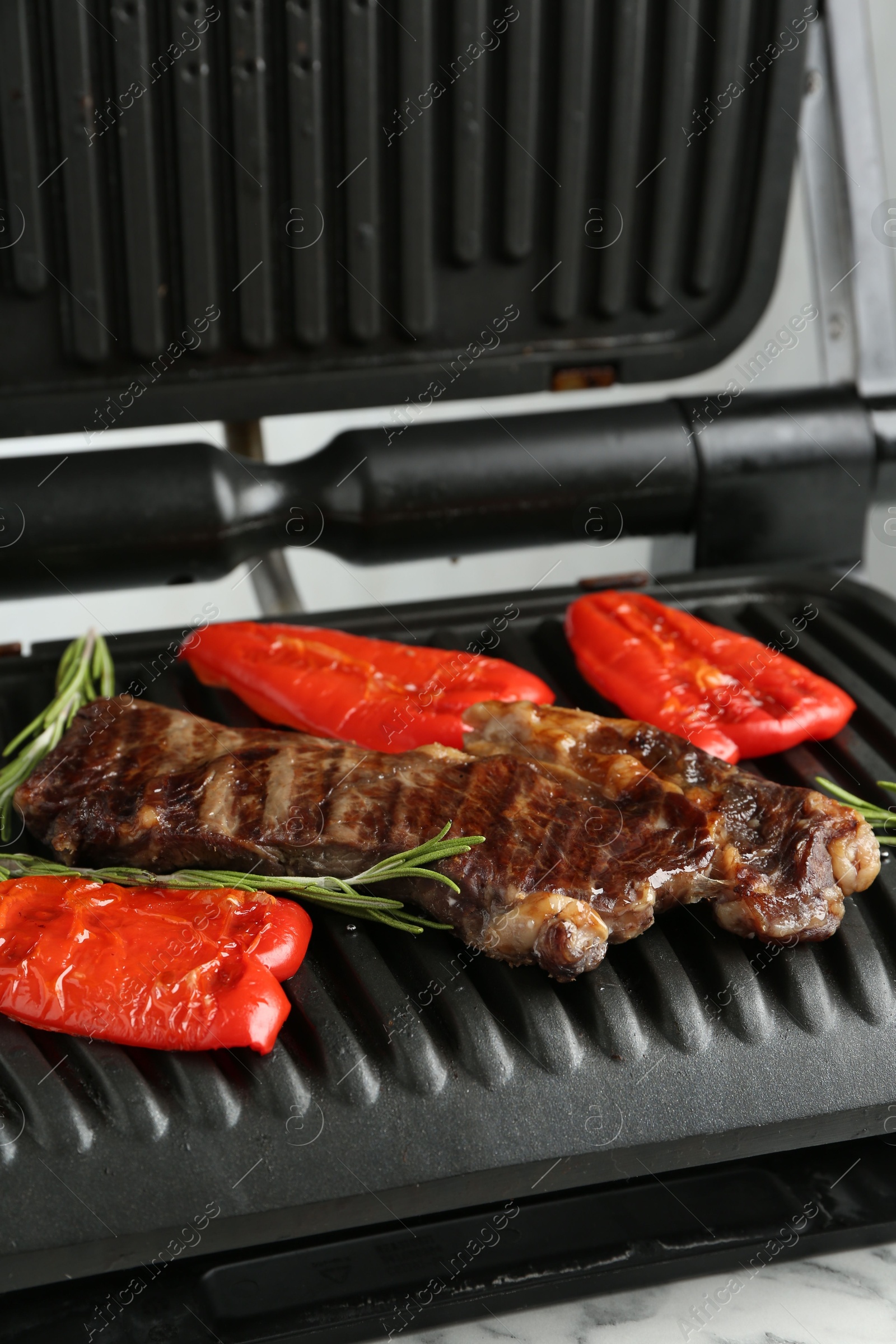 Photo of Electric grill with beef steaks, bell peppers and rosemary on white marble table, closeup