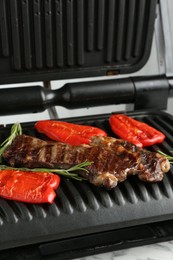 Photo of Electric grill with beef steaks, bell peppers and rosemary on white marble table, closeup