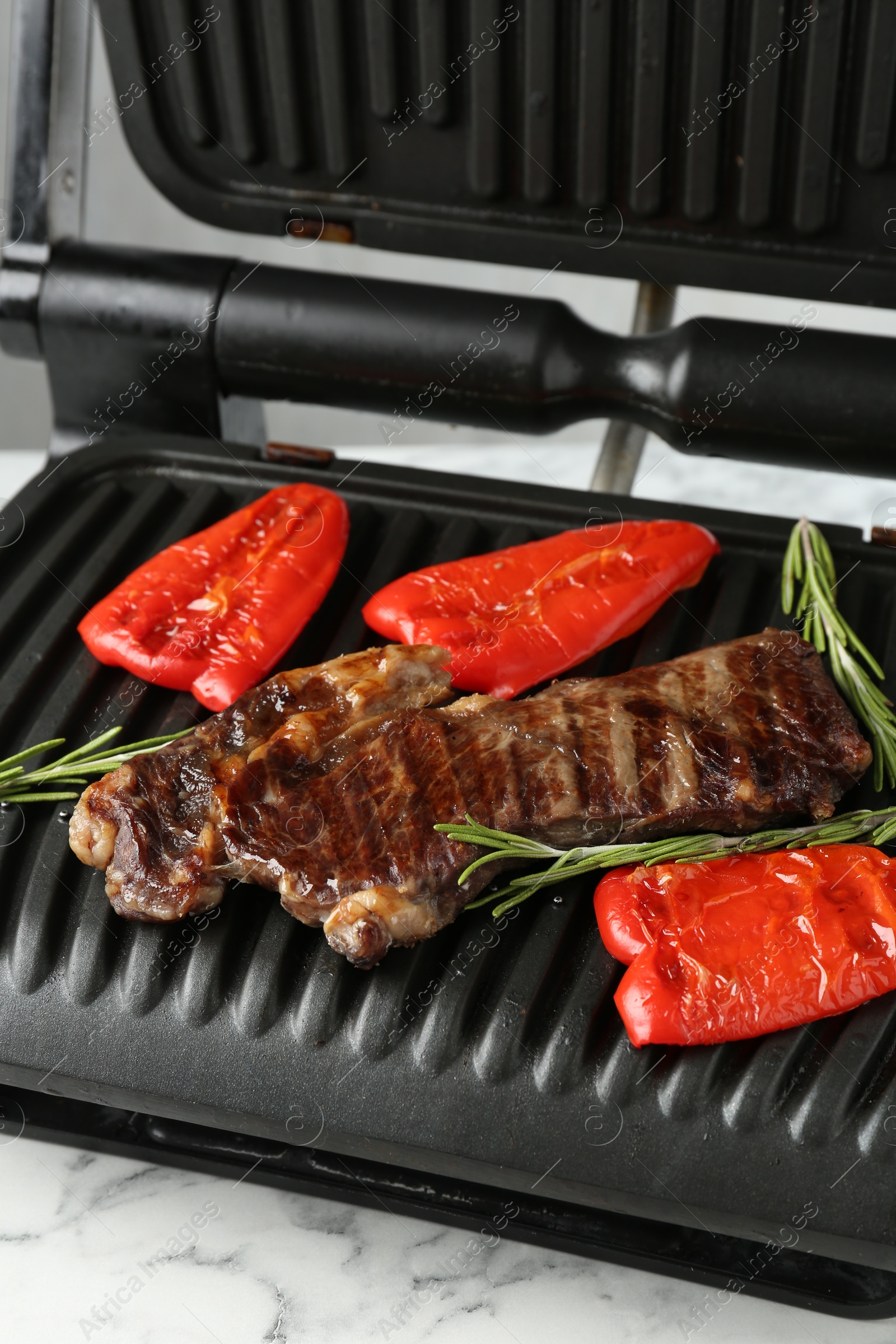 Photo of Electric grill with beef steaks, bell peppers and rosemary on white marble table, closeup