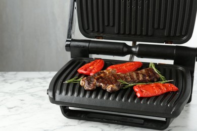 Photo of Electric grill with beef steaks, bell peppers and rosemary on white marble table, closeup. Space for text