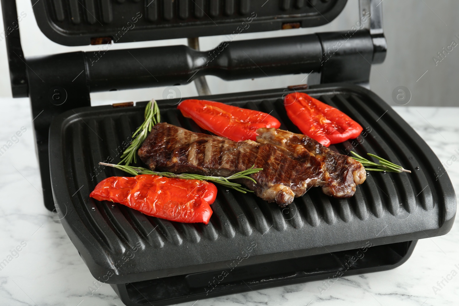 Photo of Electric grill with beef steaks, bell peppers and rosemary on white marble table, closeup