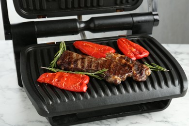 Photo of Electric grill with beef steaks, bell peppers and rosemary on white marble table, closeup