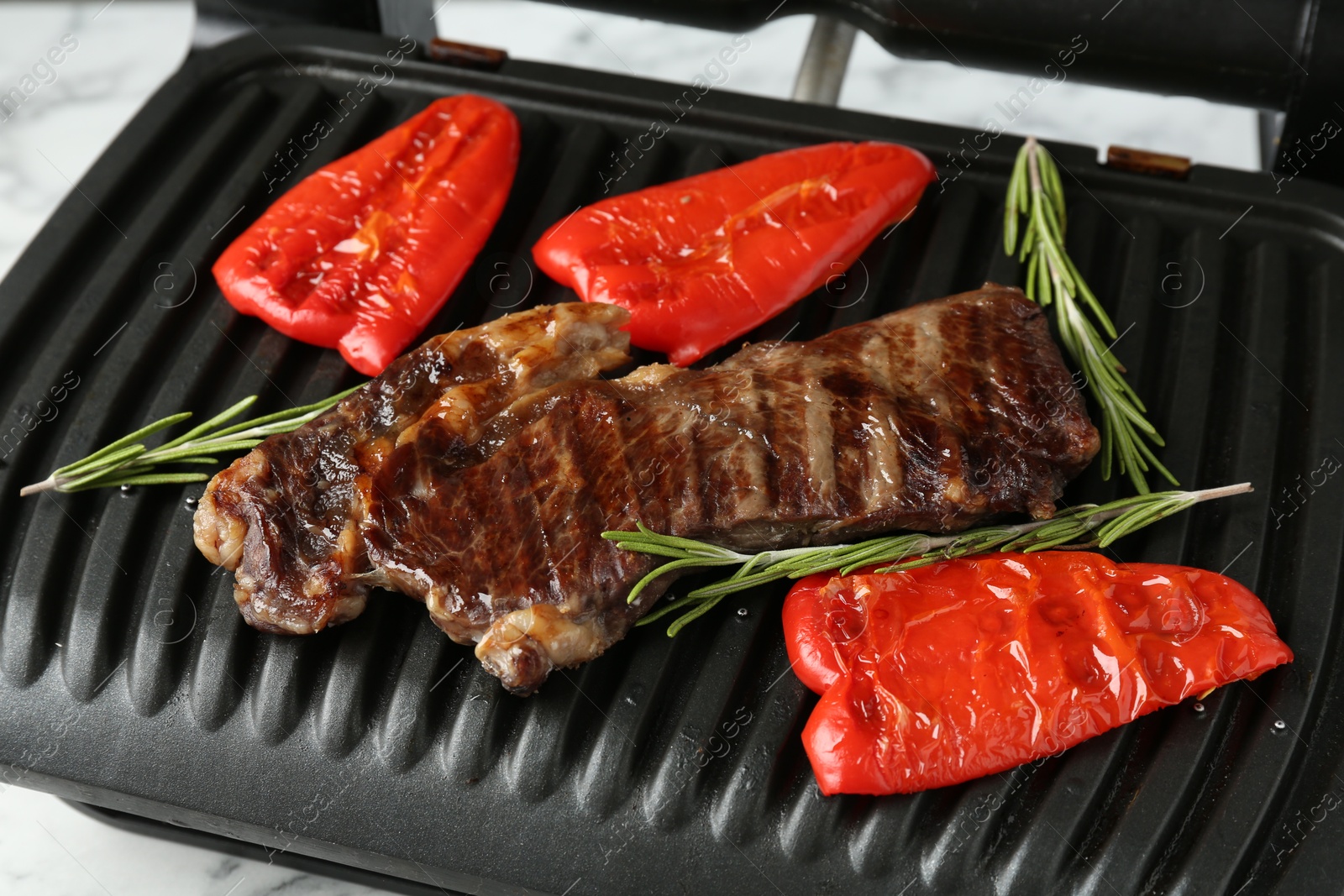 Photo of Electric grill with beef steaks, bell peppers and rosemary on white table, closeup