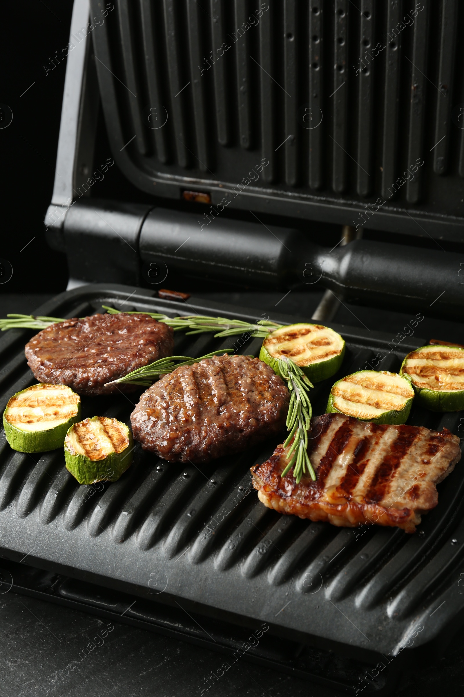 Photo of Electric grill with beef steaks, zucchini and rosemary on black table, closeup
