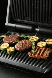 Photo of Electric grill with beef steaks, zucchini and rosemary on black table, closeup