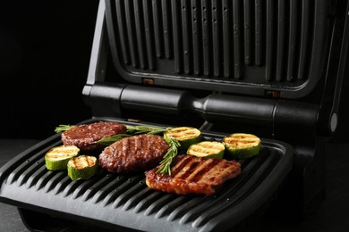 Photo of Electric grill with beef steaks, zucchini and rosemary on black table, closeup