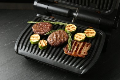 Photo of Electric grill with beef steaks, zucchini and rosemary on black table, closeup