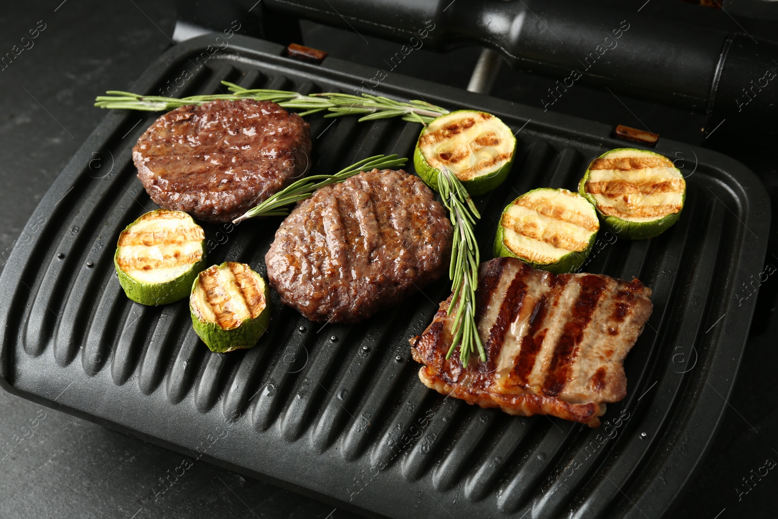 Photo of Electric grill with beef steaks, zucchini and rosemary on black table, closeup