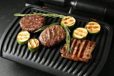 Photo of Electric grill with beef steaks, zucchini and rosemary on black table, closeup