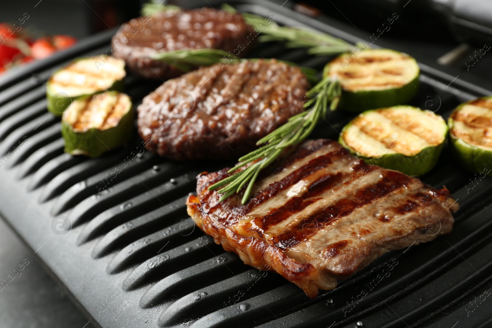 Photo of Electric grill with beef steaks, zucchini and rosemary on black table, closeup