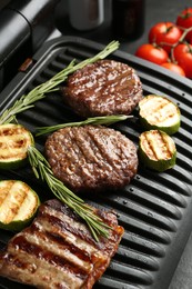 Photo of Electric grill with beef steaks, zucchini and rosemary on black table, closeup