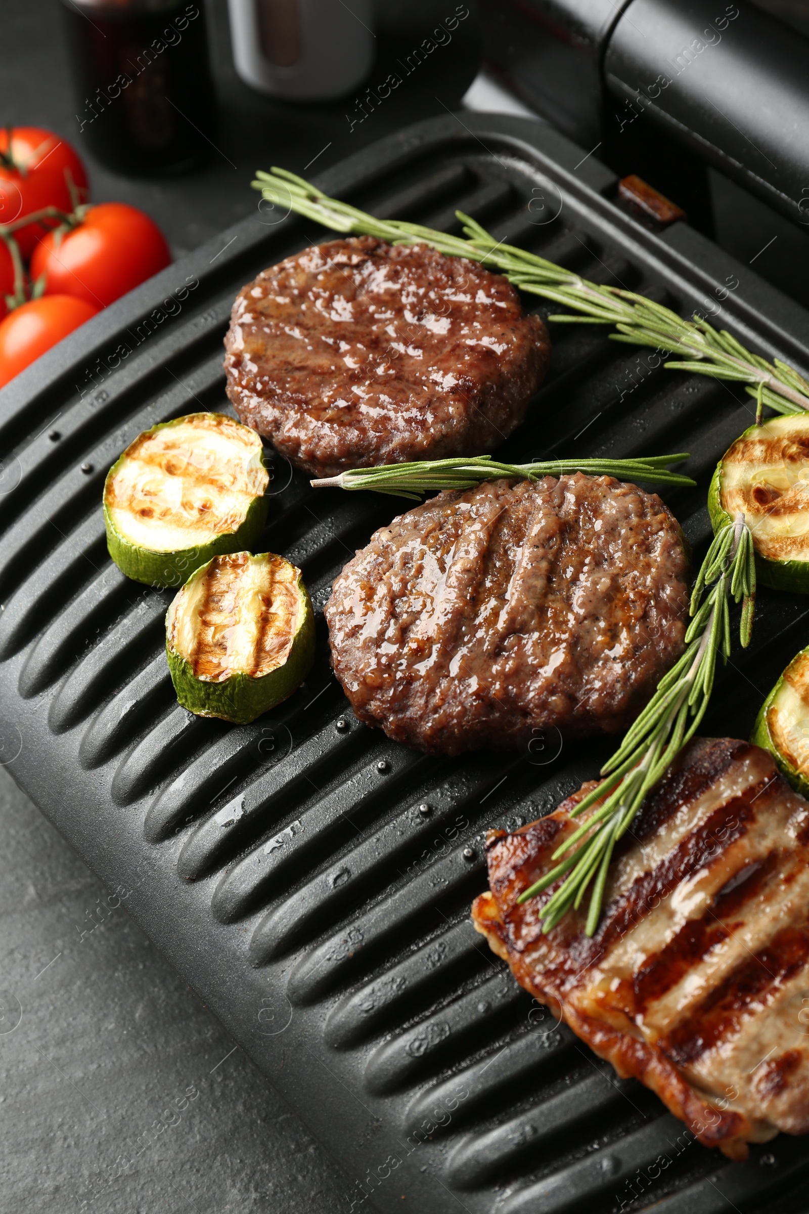 Photo of Electric grill with beef steaks, zucchini and rosemary on black table, closeup