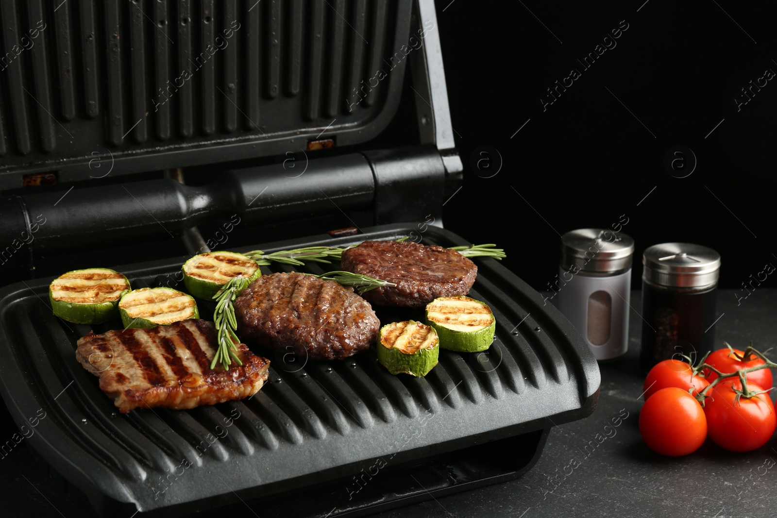 Photo of Electric grill with beef steaks, zucchini and rosemary on black table, closeup