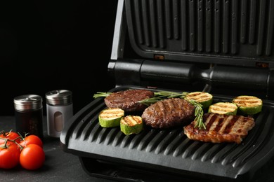Photo of Electric grill with beef steaks, zucchini and rosemary on black table, closeup