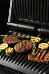 Photo of Electric grill with beef steaks, zucchini and rosemary on black table, closeup