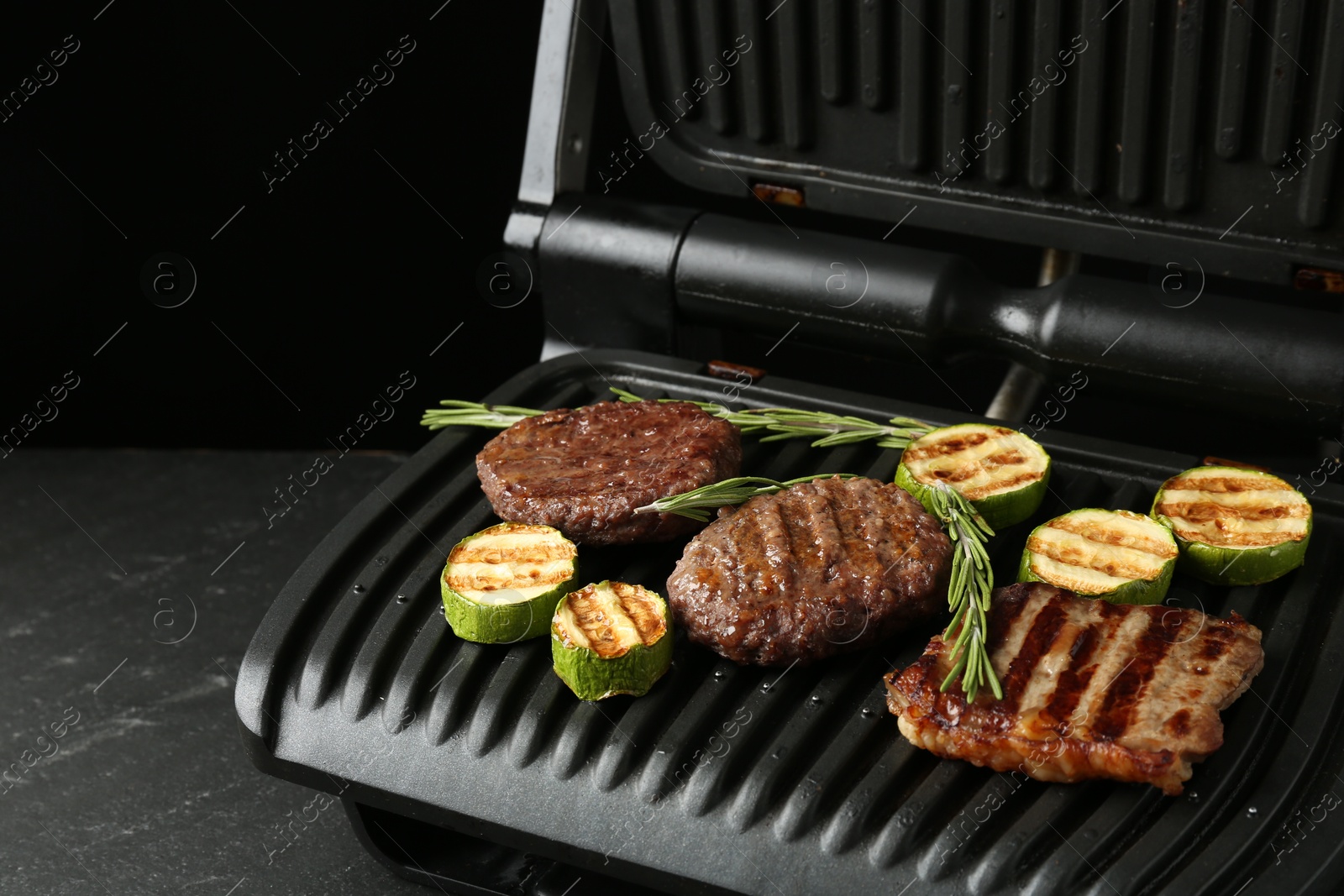 Photo of Electric grill with beef steaks, zucchini and rosemary on black table, closeup. Space for text