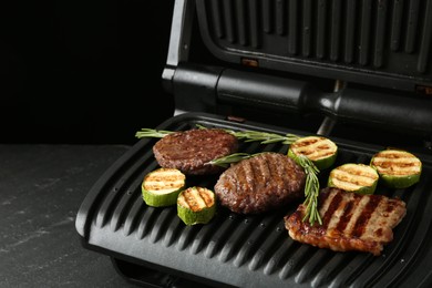 Photo of Electric grill with beef steaks, zucchini and rosemary on black table, closeup. Space for text