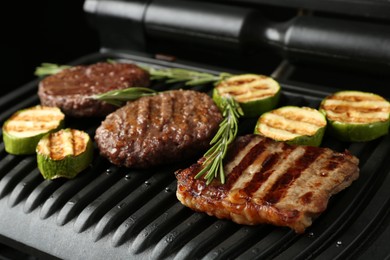 Photo of Electric grill with beef steaks, zucchini and rosemary on black table, closeup
