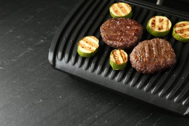 Photo of Electric grill with beef and zucchini on black table, closeup. Space for text