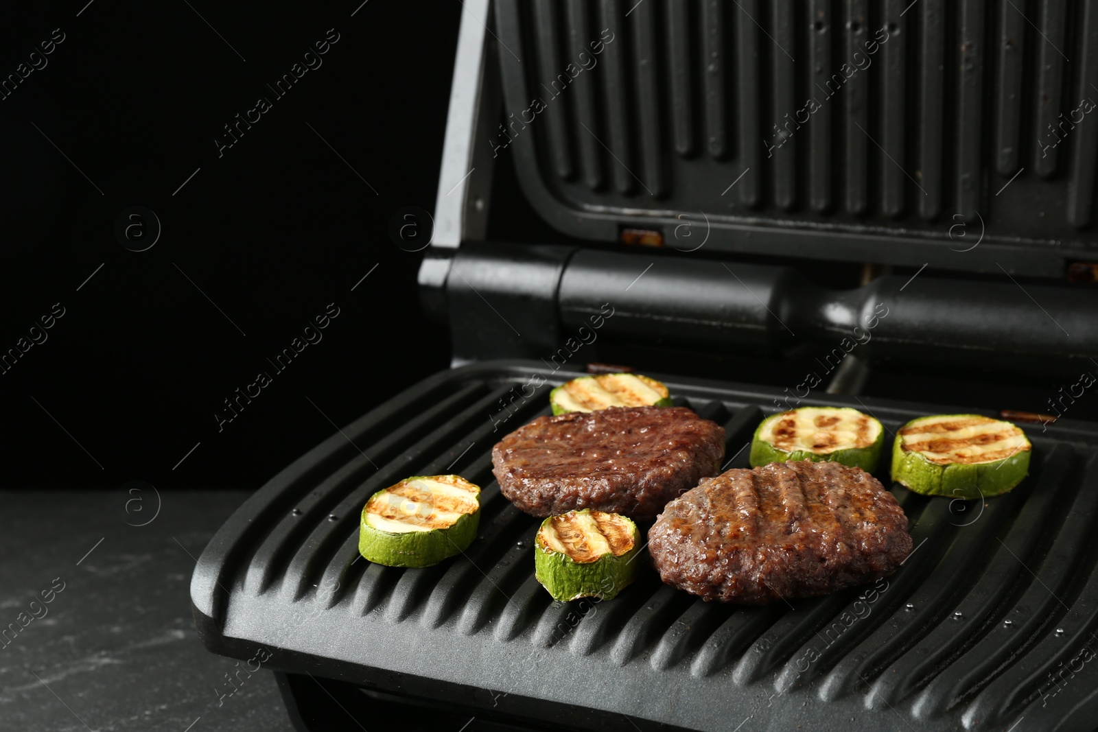 Photo of Electric grill with beef and zucchini on black table, closeup