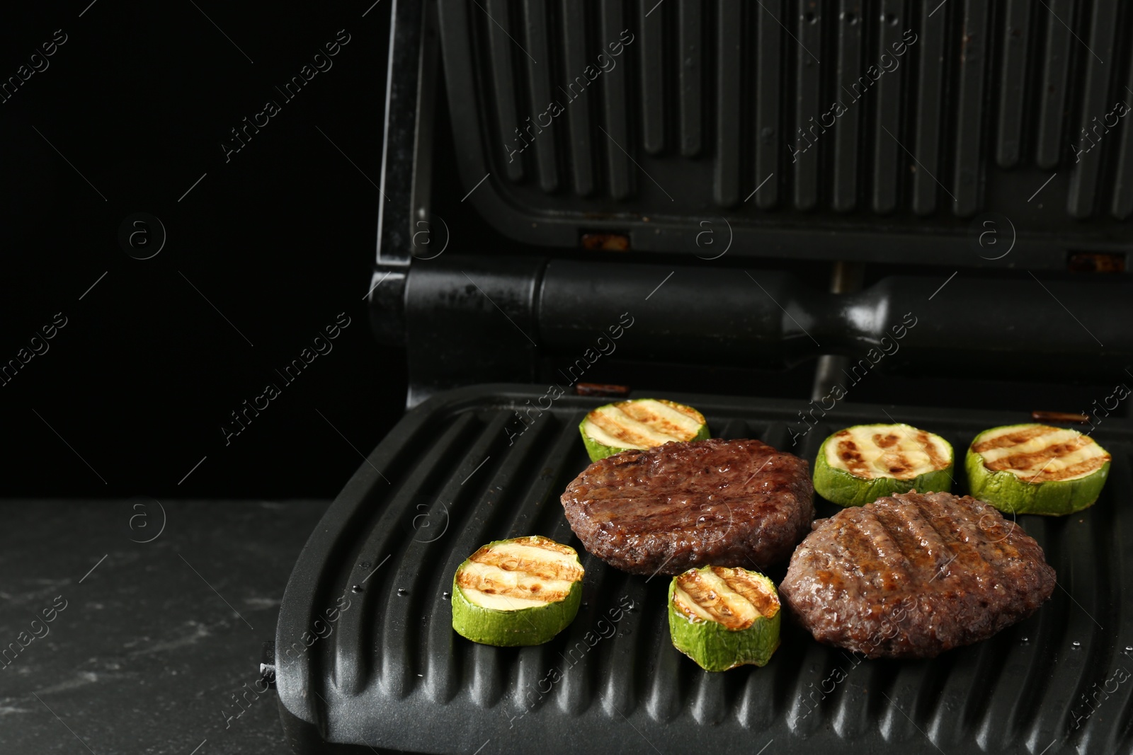 Photo of Electric grill with beef and zucchini on black table, closeup