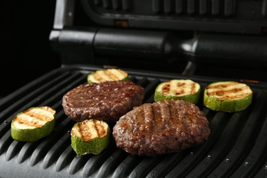 Photo of Electric grill with beef and zucchini on black table, closeup