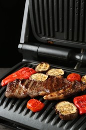 Photo of Electric grill with beef steaks and vegetables on grey table, closeup