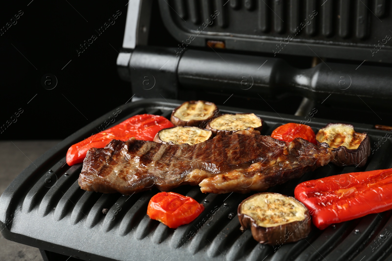 Photo of Electric grill with beef steaks and vegetables on grey table, closeup