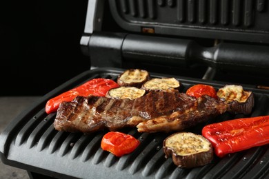 Photo of Electric grill with beef steaks and vegetables on grey table, closeup