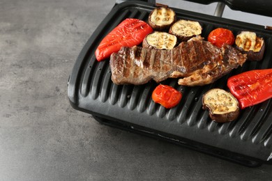 Photo of Electric grill with beef steaks and vegetables on grey table, closeup. Space for text