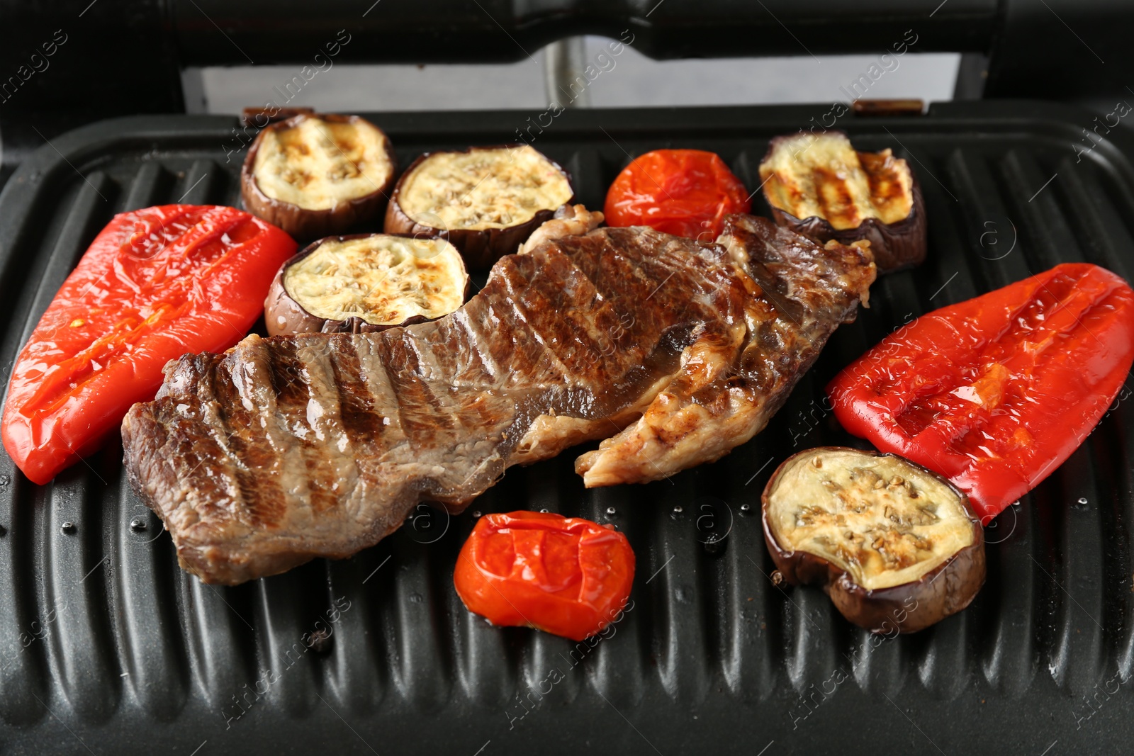 Photo of Electric grill with beef steaks and vegetables on grey table, closeup