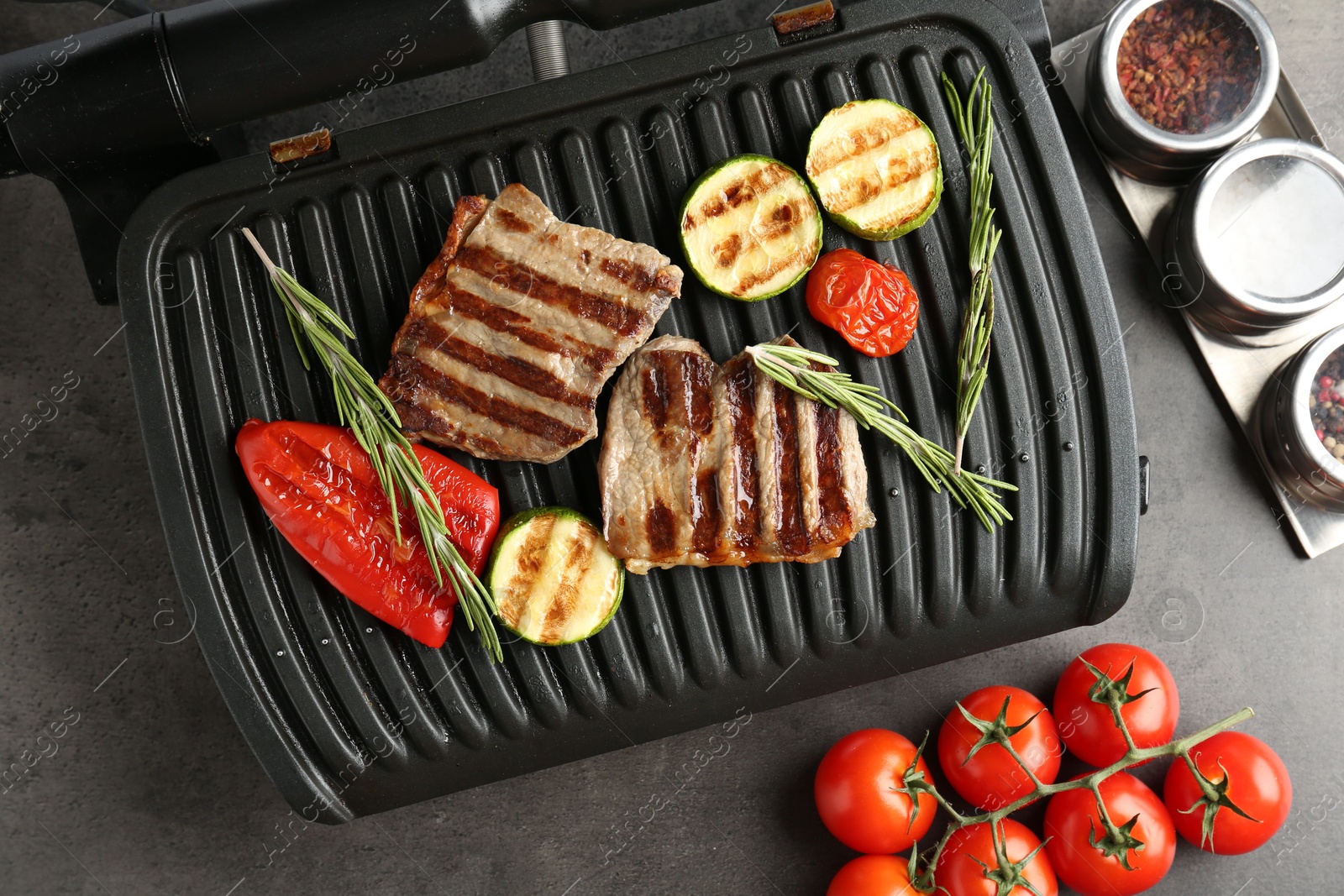 Photo of Electric grill with beef steaks, vegetables and rosemary on grey table, flat lay