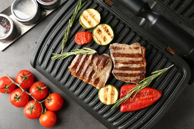 Photo of Electric grill with beef steaks, vegetables and rosemary on grey table, flat lay