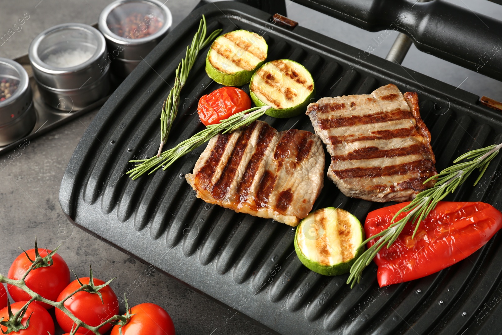 Photo of Electric grill with beef steaks, vegetables and rosemary on grey table, closeup