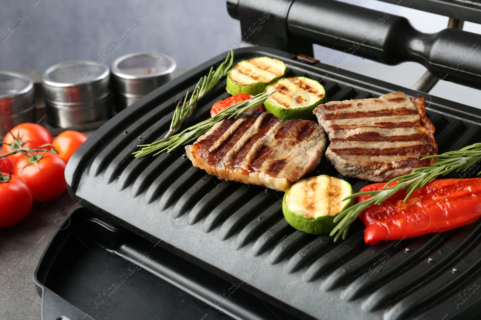 Photo of Electric grill with beef steaks, vegetables and rosemary on grey table, closeup