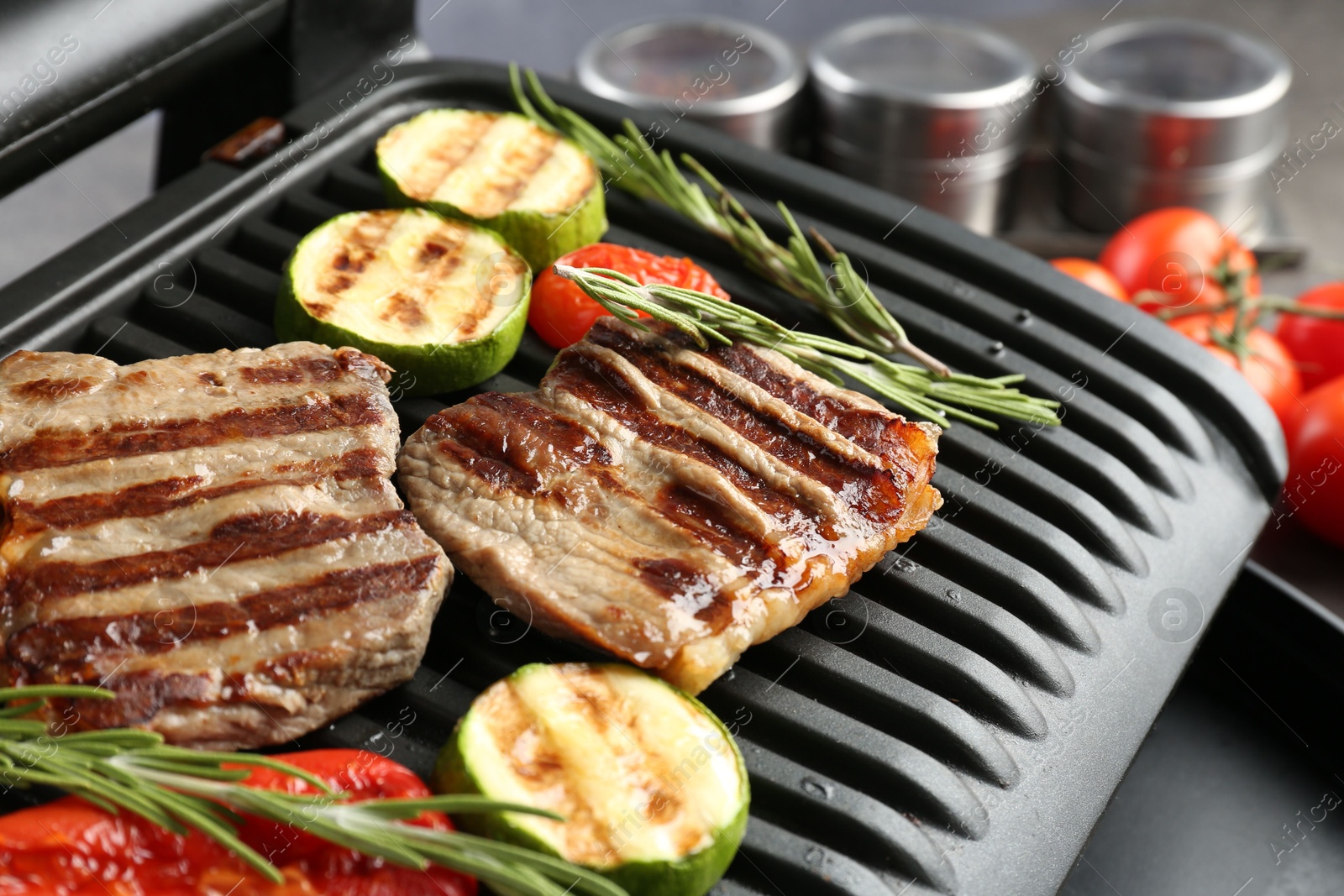 Photo of Electric grill with beef steaks, vegetables and rosemary on grey table, closeup