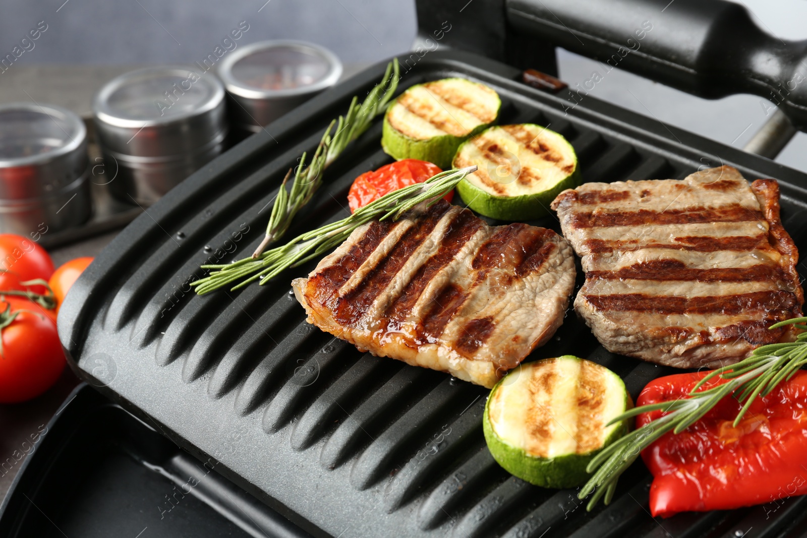 Photo of Electric grill with beef steaks, vegetables and rosemary on grey table, closeup